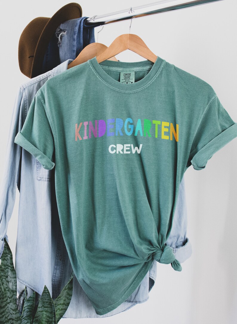 May include: A teal green t-shirt with the words "Kindergarten Crew" printed in rainbow colors. The shirt is tied in a knot and hanging on a wooden hanger.