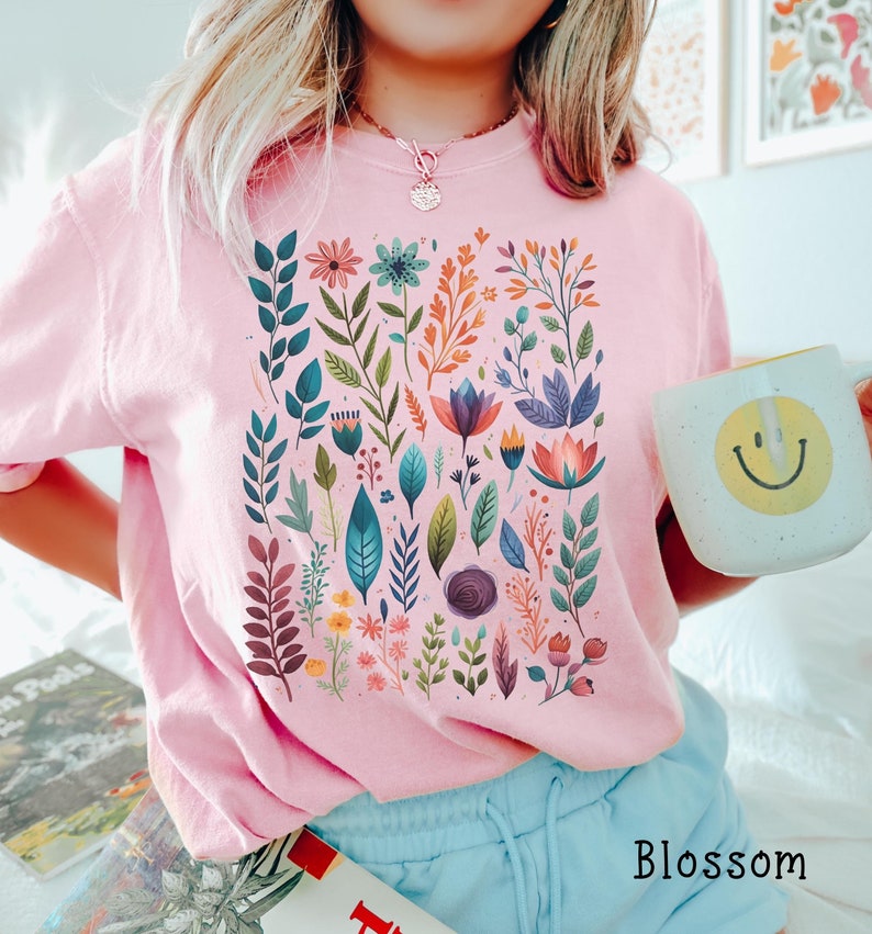 May include: A pink t-shirt with a colorful floral print featuring leaves, flowers, and stems. The shirt has a crew neck and short sleeves. The word "Blossom" is printed on the bottom right corner of the shirt.