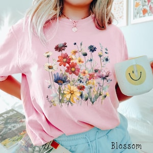 May include: A pink t-shirt with a floral print featuring a variety of colorful flowers and green leaves. The shirt has a crew neck and short sleeves. The word "Blossom" is printed on the bottom right corner of the image.