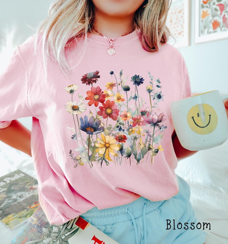 May include: A pink t-shirt with a floral print featuring a variety of colorful flowers and green leaves. The shirt has a crew neck and short sleeves. The word "Blossom" is printed on the bottom right corner of the image.