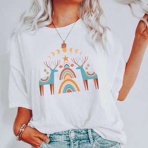 Puede incluir: Camiseta blanca con un dise&ntilde;o gr&aacute;fico de dos renos estilizados con cuernos, de pie frente a un arco&iacute;ris con una estrella en la parte superior. El dise&ntilde;o est&aacute; en tonos de azul, marr&oacute;n, naranja, amarillo y blanco.