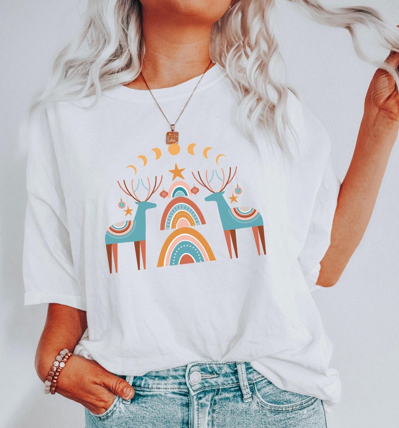 Puede incluir: Camiseta blanca con un dise&ntilde;o gr&aacute;fico de dos renos estilizados con cuernos, de pie frente a un arco&iacute;ris con una estrella en la parte superior. El dise&ntilde;o est&aacute; en tonos de azul, marr&oacute;n, naranja, amarillo y blanco.