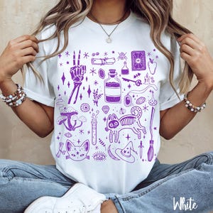 May include: White t-shirt with a purple print of various witchy and spooky illustrations, including a cat, a skeleton hand, a potion bottle, a witch's hat, and a broom.