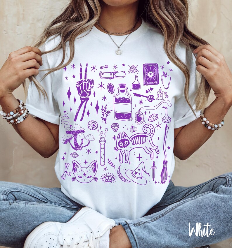 May include: White t-shirt with a purple print of various witchy and spooky illustrations, including a cat, a skeleton hand, a potion bottle, a witch's hat, and a broom.