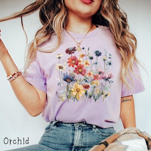 May include: A lavender t-shirt with a floral print featuring a variety of colorful flowers, including yellow, red, pink, and blue. The shirt has a relaxed fit and a crew neck.