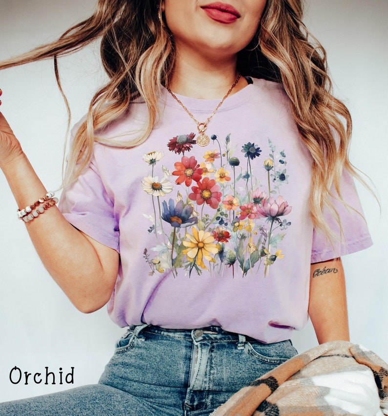 May include: A lavender t-shirt with a floral print featuring a variety of colorful flowers, including yellow, red, pink, and blue. The shirt has a relaxed fit and a crew neck.