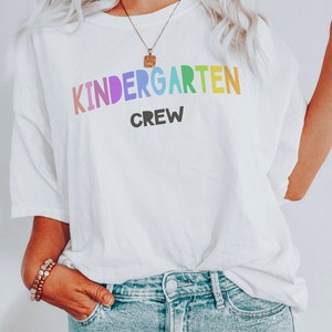 May include: White t-shirt with the text "KINDERGARTEN CREW" in rainbow colors. The word "KINDERGARTEN" is in a large, bold font, and the word "CREW" is in a smaller, black font.