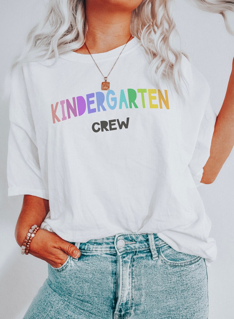May include: White t-shirt with the text "KINDERGARTEN CREW" in rainbow colors. The word "KINDERGARTEN" is in a large, bold font, and the word "CREW" is in a smaller, black font.