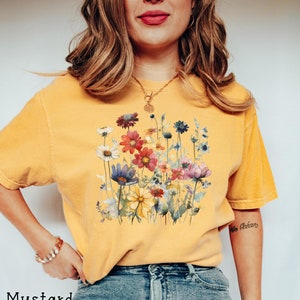 May include: A mustard yellow t-shirt with a watercolor floral print. The flowers are in shades of pink, red, yellow, blue, and purple.