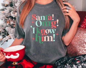 Puede incluir: Una camiseta gris con el texto "Santa!  Omg! I know him!" en letras de colores. La camiseta la lleva puesta una persona sentada en una manta de cuadros rojos y negros.