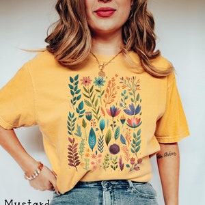 May include: A mustard yellow t-shirt with a colorful floral print. The print features a variety of flowers, leaves, and stems in shades of pink, blue, green, and orange.