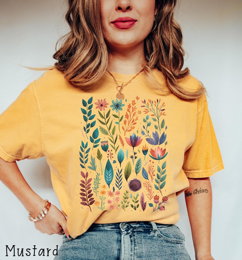May include: A mustard yellow t-shirt with a colorful floral print. The print features a variety of flowers, leaves, and stems in shades of pink, blue, green, and orange.