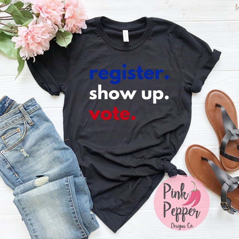 May include: Black t-shirt with the text "register. show up. vote." in blue, white, and red lettering. The shirt is laid flat on a white surface with a pair of brown sandals and blue jeans.