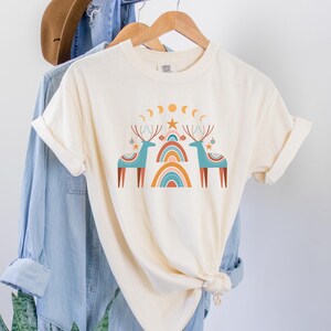 Puede incluir: Una camiseta de color crema con un dise&ntilde;o gr&aacute;fico de dos renos azules con cuernos y adornos rojos. Los renos est&aacute;n de pie frente a un arco&iacute;ris estilizado con colores naranja, rosa y azul. Hay lunas amarillas y una estrella amarilla sobre los renos.
