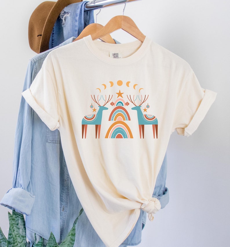 Puede incluir: Una camiseta de color crema con un dise&ntilde;o gr&aacute;fico de dos renos azules con cuernos y adornos rojos. Los renos est&aacute;n de pie frente a un arco&iacute;ris estilizado con colores naranja, rosa y azul. Hay lunas amarillas y una estrella amarilla sobre los renos.