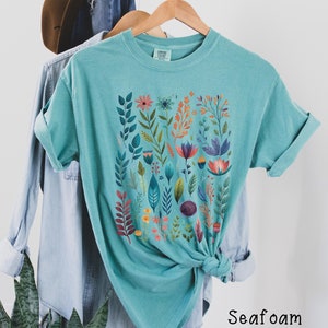 May include: A teal blue t-shirt with a colorful floral print. The print features a variety of flowers, leaves, and stems in shades of pink, orange, purple, and green. The shirt is made of a soft, comfortable fabric and has a relaxed fit.
