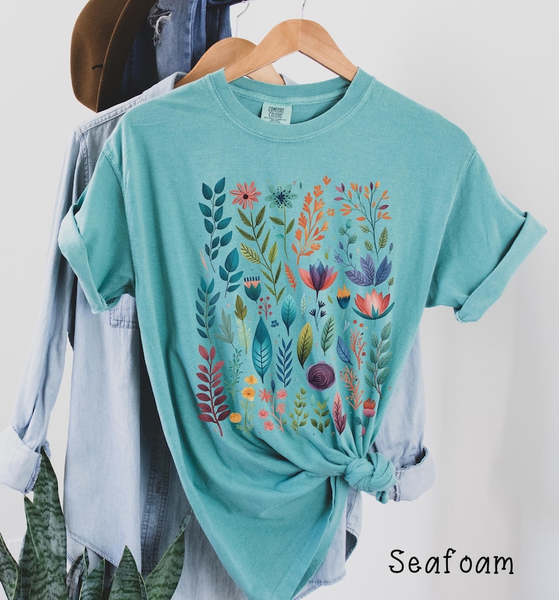 May include: A teal blue t-shirt with a colorful floral print. The print features a variety of flowers, leaves, and stems in shades of pink, orange, purple, and green. The shirt is made of a soft, comfortable fabric and has a relaxed fit.