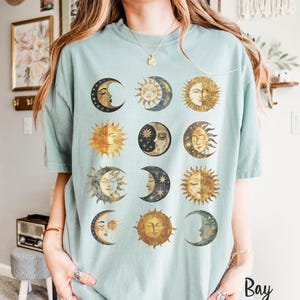 May include: A light blue t-shirt with a graphic of twelve moons and suns in a grid pattern. The moons and suns are illustrated in a vintage style with gold and brown accents.