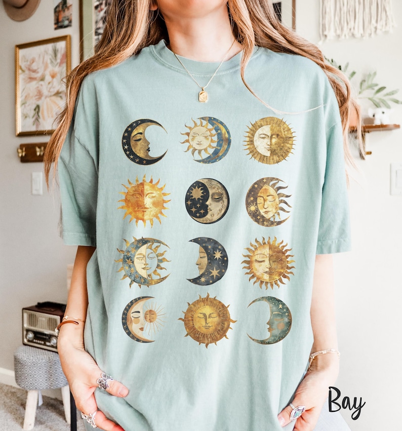 May include: A light blue t-shirt with a graphic of twelve moons and suns in a grid pattern. The moons and suns are illustrated in a vintage style with gold and brown accents.