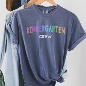 May include: A blue t-shirt with the text "Kindergarten Crew" in rainbow colors. The shirt is hanging on a wooden hanger.