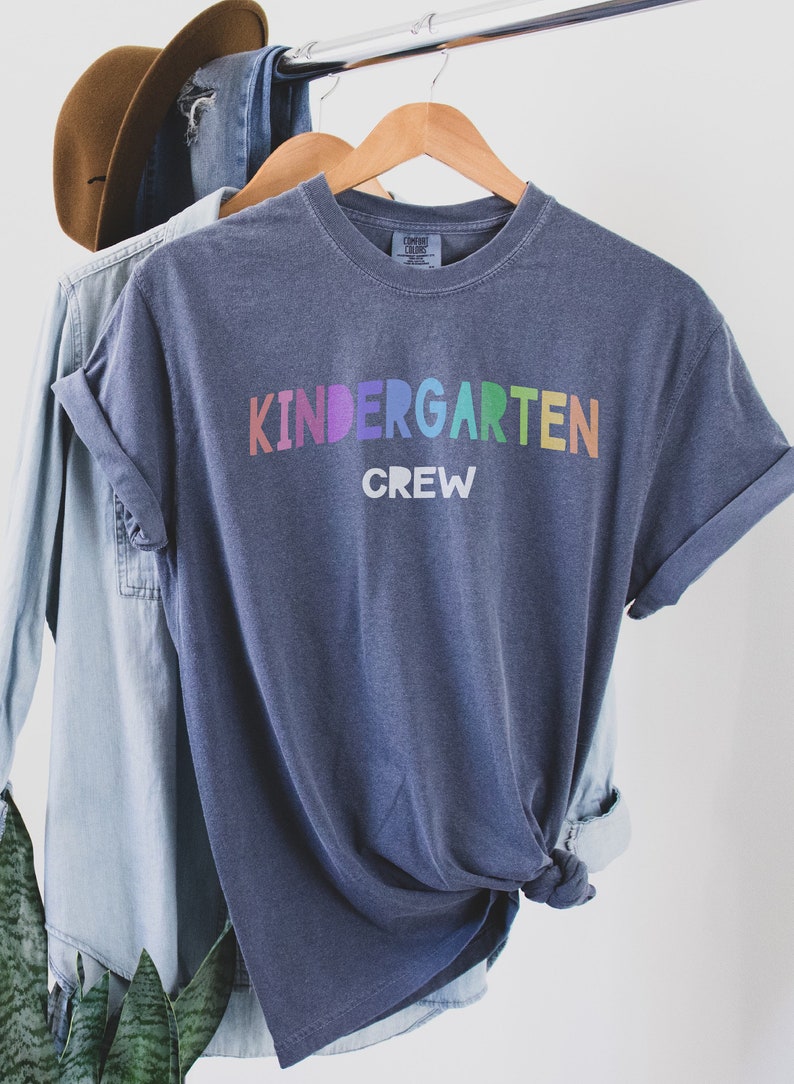 May include: A blue t-shirt with the text "Kindergarten Crew" in rainbow colors. The shirt is hanging on a wooden hanger.