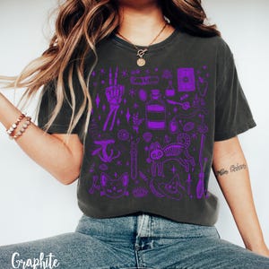 May include: A dark gray t-shirt with a purple graphic print of various witchy items, including a cat, a skeleton, a potion bottle, a hand with a peace sign, a broom, a hat, and a tarot card. The shirt has a crew neck and short sleeves.