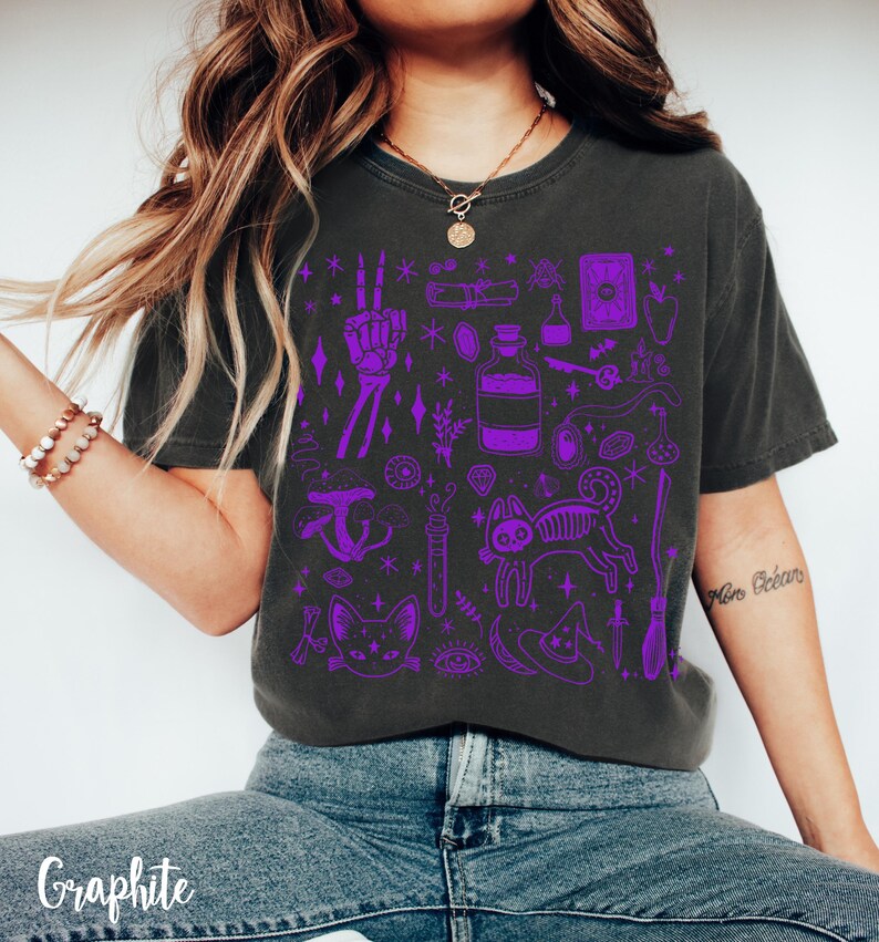 May include: A dark gray t-shirt with a purple graphic print of various witchy items, including a cat, a skeleton, a potion bottle, a hand with a peace sign, a broom, a hat, and a tarot card. The shirt has a crew neck and short sleeves.