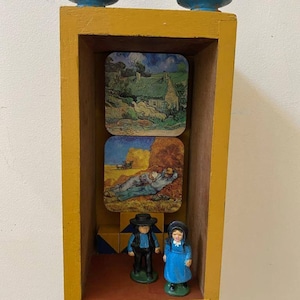 May include: A yellow wooden box with a glass front, containing three square coasters with images of a house, a field of wheat, and a man sleeping. Two small figurines of people in blue and black clothing stand in front of the coasters.