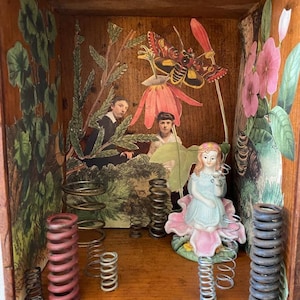 May include: A wooden box with a collage of images and objects. The box features a butterfly, flowers, a girl figurine, and several metal springs. The box is a mixed media art piece.