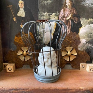May include: A white porcelain doll's head is encased in a wire cage, sitting on a wooden shelf. The doll's head is surrounded by gold and brown metal accents. The shelf is part of a larger wooden structure. The background features two religious figures in a painting.