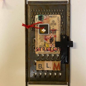 May include: A metal grater with a red string attached to the top. A card with the word "STRETCH" is attached to the grater. There are four wooden blocks spelling out "BLM" on the grater. A black cross is attached to the right side of the grater.