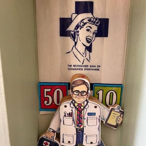 May include: A wooden toy doctor figure wearing a white coat, a red tie, and glasses. The figure is holding a clipboard in one hand and a red medical bag in the other. The figure is standing in front of a white sign with a blue cross and the text "The recognized sign of cleanliness everywhere."