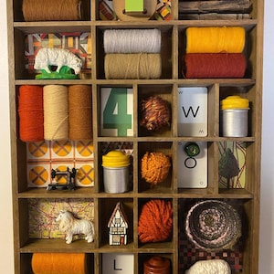 May include: A wooden shadow box with 16 compartments filled with various objects, including yarn, fabric scraps, toy sheep, and small containers. The box has a rustic, vintage aesthetic.