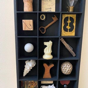 May include: A black shadow box with 12 compartments, each containing a different object. The objects include a cork, a ticket, a lightbulb, a key, a number 8, a fishing hook, a white ball, a chess piece, a seashell, a wooden Y, a golf ball, a postcard, a cow figurine, and a chess piece.