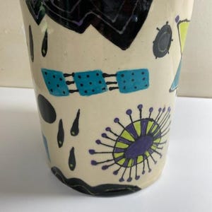 May include: A white ceramic planter with a black, blue, green, and purple abstract design. The planter has a rounded shape and a wide opening.