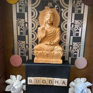 Könnte beinhalten: Eine Holzstatue von Buddha, der in meditativer Haltung auf einer Lotusblume sitzt. Die Statue steht auf einer schwarzen Box mit dem Wort "BUDDHA" in Holzbuchstaben. Der Hintergrund ist eine schwarz-goldene Tapete.