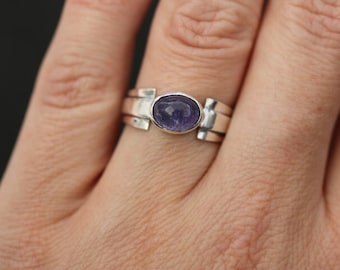 May include: A silver ring featuring a purple gemstone set in a bezel setting. The ring has a simple, modern design and is crafted from sterling silver.
