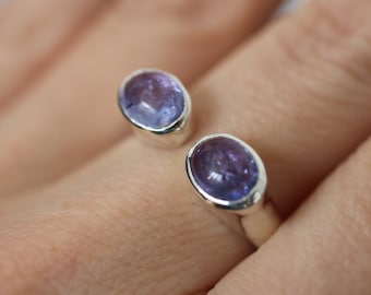 May include: A silver ring with two oval-shaped purple gemstones. The ring is open and sits on a finger.