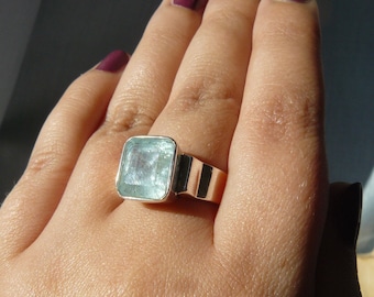 May include: A silver ring with a large, light blue aquamarine gemstone. The ring is set in a simple, square-shaped band.