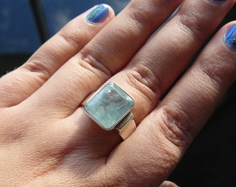 May include: A silver ring with a large, light blue, square-shaped gemstone. The ring is on a person's finger.