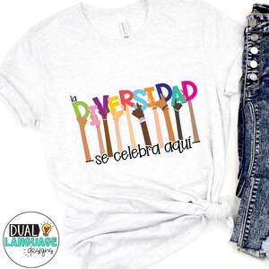 May include: A light grey t-shirt with the words "la DIVERSIDAD se celebra aqu&iacute;" in colourful letters. The design features diverse hands raised in the air, celebrating inclusivity. The shirt is made of soft material.