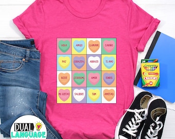 May include: Pink t-shirt featuring a graphic of a grid of hearts with Spanish and English words. The words are "Hola", "Amigo", "Llámame", "Cariño", "Paz", "Corazón", "Abrazo", "Te Amo", "Besos", "Escribime", "Amor", "Sonrie", "Me Gustas", "Saludos", "TQM", "Amistad".