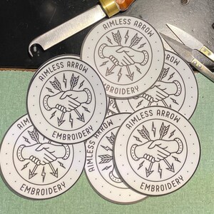 Puede incluir: Cinco pegatinas redondas plateadas con el texto "Aimless Arrow Embroidery" y un gráfico de dos manos dándose la mano con rayos alrededor.