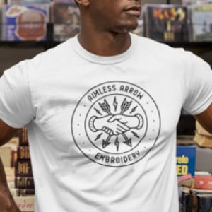 Puede incluir: Camiseta blanca de cuello redondo con un gráfico circular en blanco y negro que dice "Aimless Arrow Embroidery". El diseño presenta dos manos estrechándose con flechas y rayos. La persona lleva gafas de sol.