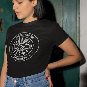 Puede incluir: Una camiseta negra con un gráfico circular blanco que presenta un apretón de manos con flechas y el texto "Rimless Arrow Embroidery".