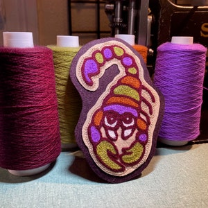 Puede incluir: Un parche bordado de escorpión colorido con hilo morado, naranja, amarillo y verde. El parche está sobre un fondo de fieltro morado oscuro. El escorpión está rodeado de carretes de hilo de varios colores.