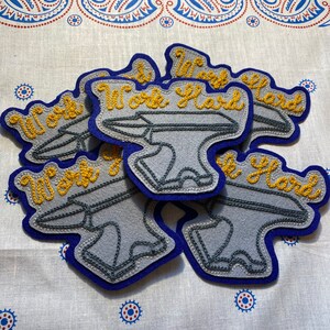 Puede incluir: Cuatro parches de fieltro gris con borde azul, cada uno con un yunque bordado y el texto "Work Hard".