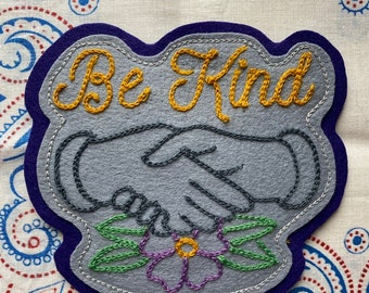 May include: A gray felt patch with a blue border, featuring a handshake with a flower and leaves below. The text "Be Kind" is embroidered in yellow thread.