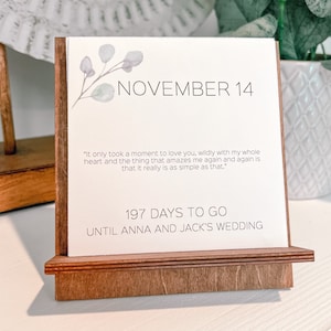 K&ouml;nnte beinhalten: Ein Holz-Schreibtischkalender zeigt "NOVEMBER 14" mit einem floralen Design. Der Kalender enth&auml;lt ein Zitat und den Text "197 DAYS TO GO UNTIL ANNA AND JACK'S WEDDING". Der Kalender ist aus hellbraunem Holz.