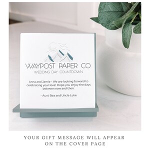 K&ouml;nnte beinhalten: Eine wei&szlig;e Karte mit blauen Bergen und dem Text "WAYPOST PAPER CO. WEDDING DAY COUNTDOWN" und einer Nachricht f&uuml;r ein Hochzeitspaar von Tante Bea und Onkel Luke. Die Karte steht auf einem hellblauen St&auml;nder. Der Text "YOUR GIFT MESSAGE WILL APPEAR ON THE COVER PAGE" befindet sich unter der Karte.